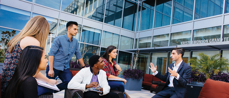 A small group of students taking notes and listening to one person talk while outside on a patio talking.