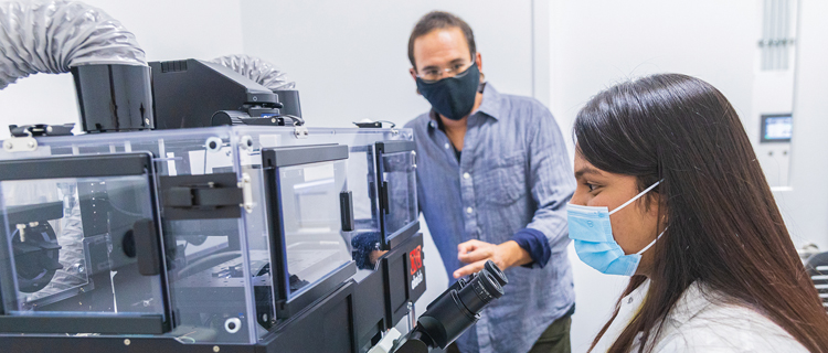 A man and a woman wearing masks with the woman in a lab coat diligently operating a machine in a laboratory at the University of California.