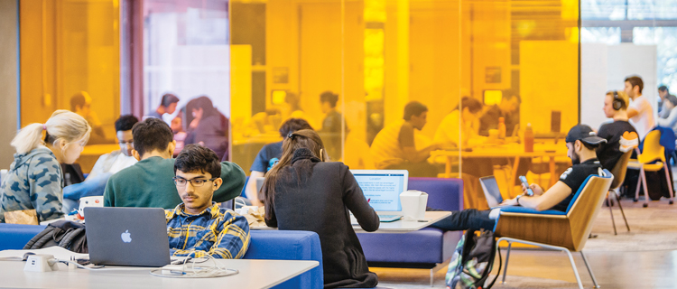 Groups of students sitting at tables working on their laptops in a spacious open room.